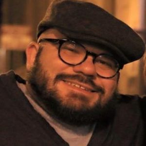 A portrait photo of Joe Stramondo. He is a white man with a brown beard wearing glasses, a brown cap, and brown v-neck sweater with a t-shirt underneathand smiling. 